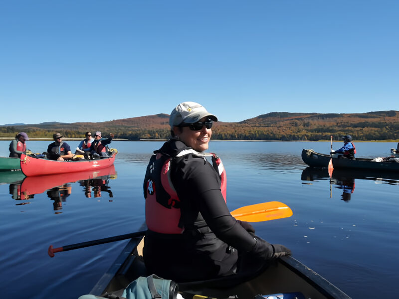 A woman in a canoe smiles at the camera, holding a paddle. She wears a grey hat, sunglasses, and a red life vest. Other canoes with people are visible in the background, along with forested hills under a clear blue sky. The water is calm and reflects the sky and surrounding landscape.